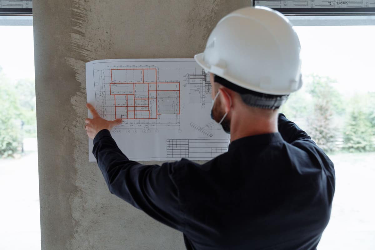 A person in a white hard hat checking a floor plan pinned to a concrete wall